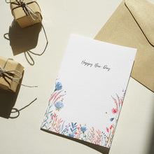 Load image into Gallery viewer, A white greeting card with 'Happy Bee Day' in script and a watercolor floral border at the bottom, shown with a kraft paper envelope on a light surface. Two small wrapped gift boxes with twine are placed nearby in a styled flat-lay arrangement.
