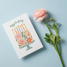 Load image into Gallery viewer, A square birthday greeting card with a colorful illustrated birthday cake and the words 'Happy Birthday' in green script, styled on a light blue background next to a pink ranunculus flower.
