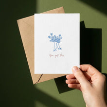 Load image into Gallery viewer, A hand holds a white greeting card with a blue line drawing of a person carrying flowers and the words 'You got this.' in red script, layered over a brown kraft envelope against a dark olive green background with strong sunlight and shadows.
